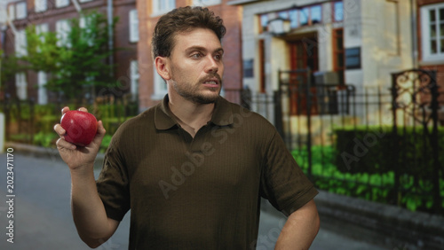 Foto Man holding red apple in his hand near a building entrance on a residential street while glancing down at the fruit; skepticism health choice