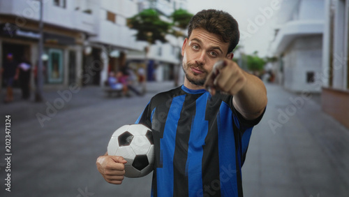 Konstfotografi Man holding a soccer ball under his arm on a city street wearing blue striped jersey and gesturing with one hand; indifference