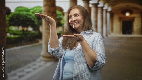 Fotografie Woman holding palms up and smiling, showing empty hands in a stone colonnade building walkway with archways and trees; joy