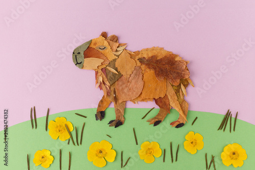 Konstfotografi Capybara made from dried autumn leaves standing on green paper grass with yellow flowers on pink background