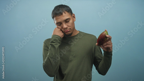 Slika na platnu Man in olive long sleeve shirt resting cheek on closed fist, holding a ripe mango in front of a soft blue studio backdrop; melancholy reflection