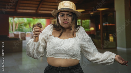 Fotografi Plus size woman holding chocolate ice cream cone, pointing finger forward, exposed stomach visible, wearing straw hat and white blouse in building lobby; annoyance refusal