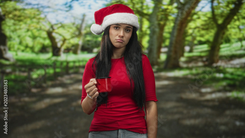 Photography Young hispanic woman in a santa hat and red shirt holds a red enamel mug with her right hand, pouting and looking at camera on a tree-lined forest path; melancholy
