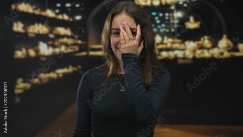 Photos Young hispanic woman with hand covering face and smiling in museum building, wearing heart pendant and dark long sleeve top; shyness sincerity
