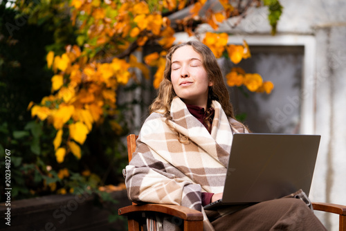 A young woman is resting her eyes after working on a laptop on a cozy autumn terrace in the daylight. The front view portrait.