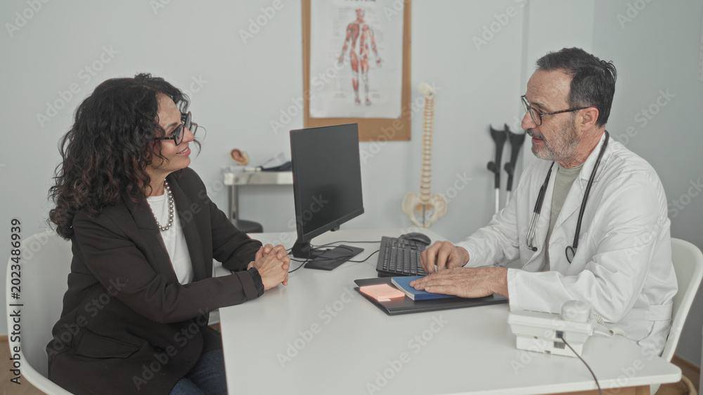 Fototapeta premium Woman patient seated with hands clasped across desk with doctor wearing stethoscope by computer and spine model in building; reassurance patient support.