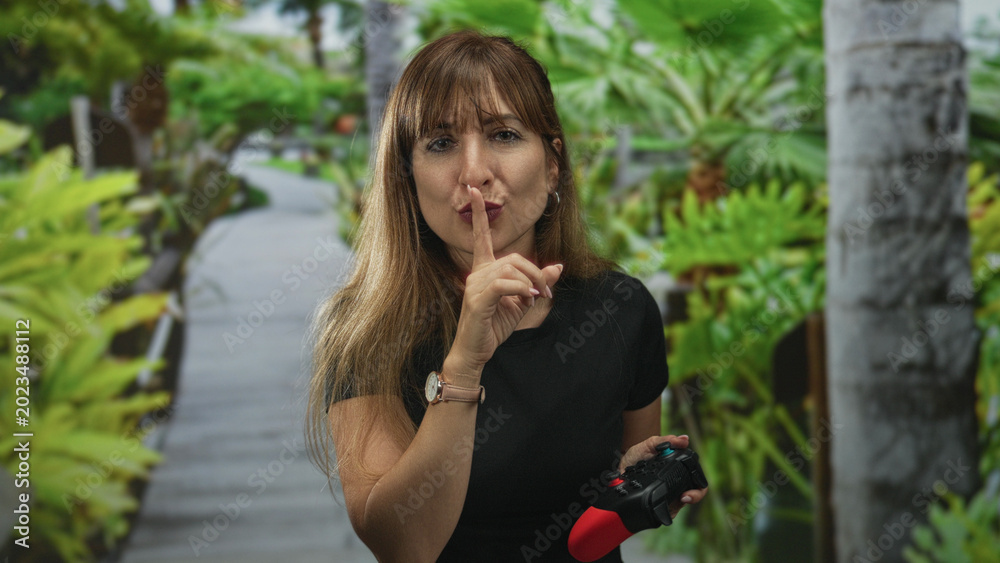 Fototapeta premium Woman holding a game controller and finger to lips for silence in forest; playful gentle secrecy hush.