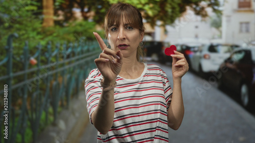 Foto Woman holding small red felt heart points finger toward camera on street; love plea determination compassion
