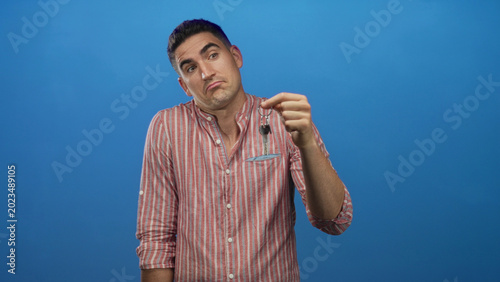 Foto Young hispanic man holding keys and shrugging in studio with solid blue backdrop; indifference dilemma