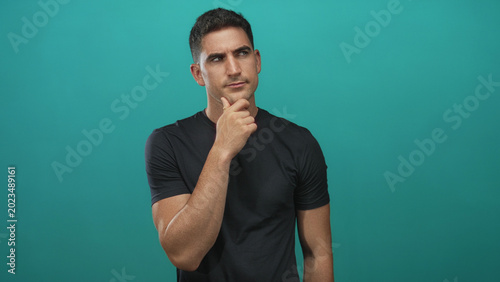 Φωτογραφία Man with hand on chin in studio with teal backdrop, wearing black tshirt and looking to the side; thoughtful reflection