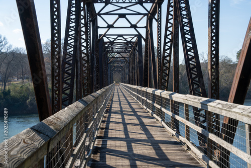 Old steel truss bridge spans calm river. Wooden walkway runs beneath iron framework. Sunlight casts long shadows across planks. Rust-colored beams rise into clear blue sky