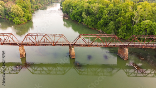 Rusty bridge spans calm, green river. Lush trees line both riverbanks. Reflections ripple gently below. Industrial decay meets natural beauty. Ideal for travel, nature, or nostalgia themes