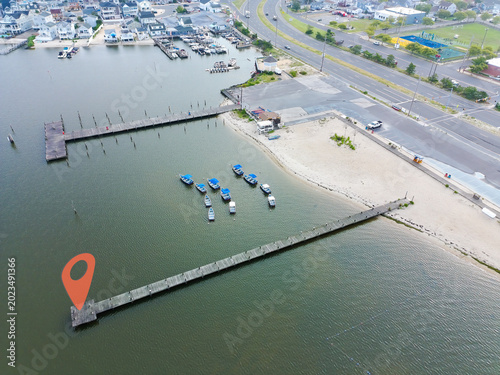 Aerial view of a harbor with boats docked at a pier. Small blue boats float near a stone-lined shore. Water reflects the sky and surrounding buildings. Road runs parallel to the dock