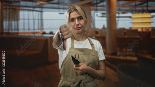 Canvas Print Woman gardener wearing apron holding a hand trowel and showing thumbs down with outstretched hand inside a dining building, direct gaze at camera; disapproval