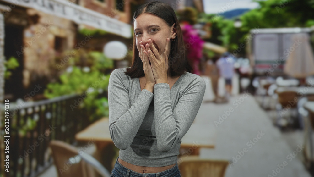 Fototapeta premium Young woman touches cheeks with both hands and smiles in a street cafe by tables and railing; spontaneous joy.