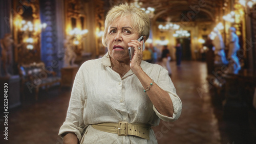 Photos Senior hispanic woman holds smartphone, frowning while checking screen and later answering a call inside an ornate building; concern