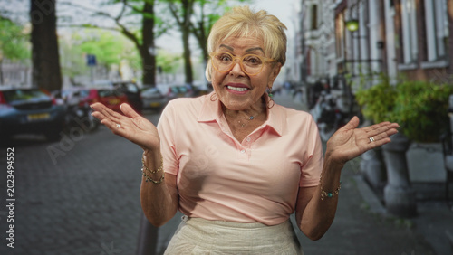 Fotografie Senior woman shrugs with palms up, wearing pink polo and glasses on a city street with parked cars and trees; bemused reflection