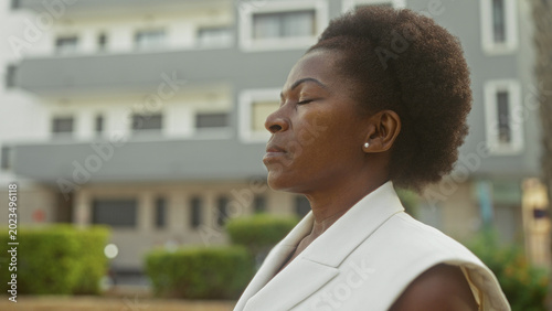 Tableau sur toile Woman with closed eyes tilting head upward, showing earring and neck, standing near modern building facade; calm reflection