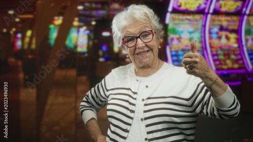 Fotografía Elderly woman raises fist at a casino slot machine, smiling with glasses, striped cardigan, ring and bracelet visible; victory luck celebration