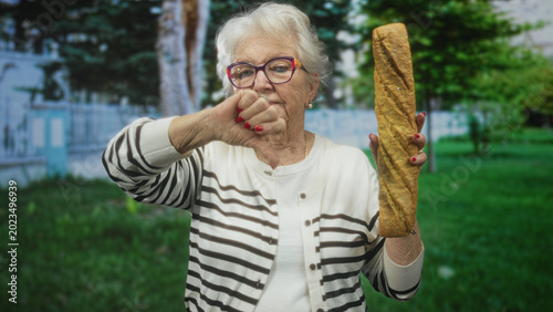 Photography Elderly woman holds a long baguette and displays thumb down gesture beside a building in a park setting; disapproval nostalgia