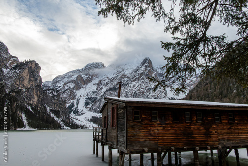 Foto A stunning view of the historic stilt boathouse over the frozen ice of Lake Braies during winter, with dense pine forests and the snowy peaks of the Dolomites under a dramatic cloudy sky