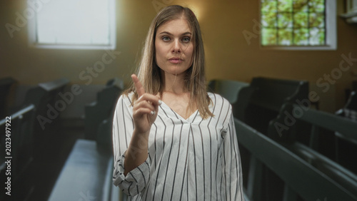 Photos Young blonde woman in striped blouse pointing finger toward wooden pews inside church building; reverence