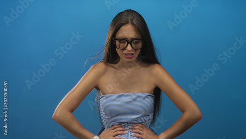 Foto Young hispanic woman clutching stomach with both hands, wearing large glasses and a strapless dress against a blue backdrop in studio; pain