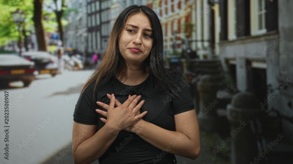 Fototapeta premium Woman with hands clasping over chest on street in city, interlaced fingers and teary expression amid urban storefronts and blurred cars; gratitude.