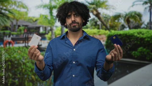 Foto Man holding two creditcards in both hands at a building pool area showing a hesitant shrug; financial uncertainty