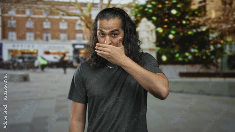 Fototapeta premium Man covering face with hand on a street plaza in front of a decorated christmas tree and statue; shame privacy.