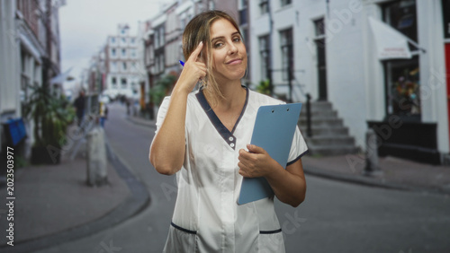 Tableau sur toile Woman nurse holding clipboard points finger to temple on a street near a building, wearing scrubs and smiling; compassionate care thoughtful