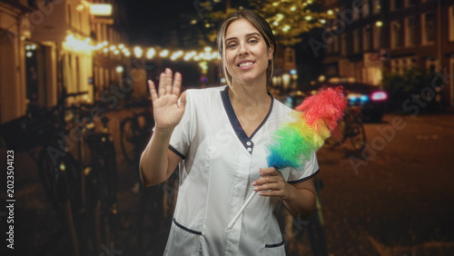 Photos Woman in a white cleaner uniform holding a rainbow feather duster and waving hand on a lit street at night; friendly greeting cheerful