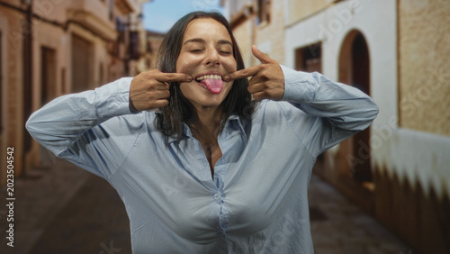 Foto Woman pulls cheeks with fingers and sticks tongue out on a street in an old town; playful mischief moment