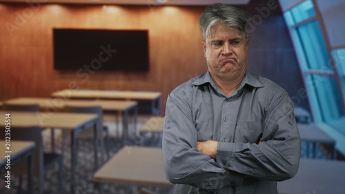 Photos Senior man arms crossed with pronounced frown standing before rows of desks and a blank monitor in a classroom; frustration concern