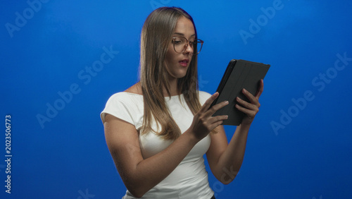 Quadro em tela Young hispanic brunette woman wearing glasses and white t shirt holding a tablet in a folio case doing a fist pump in a studio; confidence success achievement