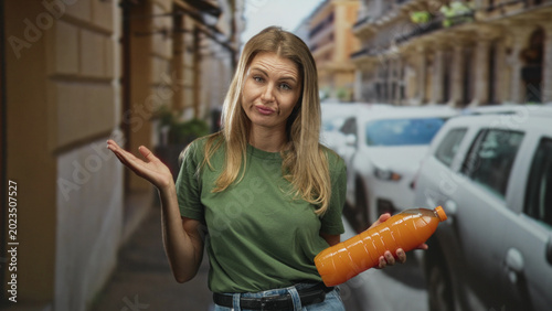 Konstfotografi Blonde woman in green shirt holding a plastic orange juice bottle in left hand and shrugging right hand among parked cars on a street; confusion uncertainty annoyance