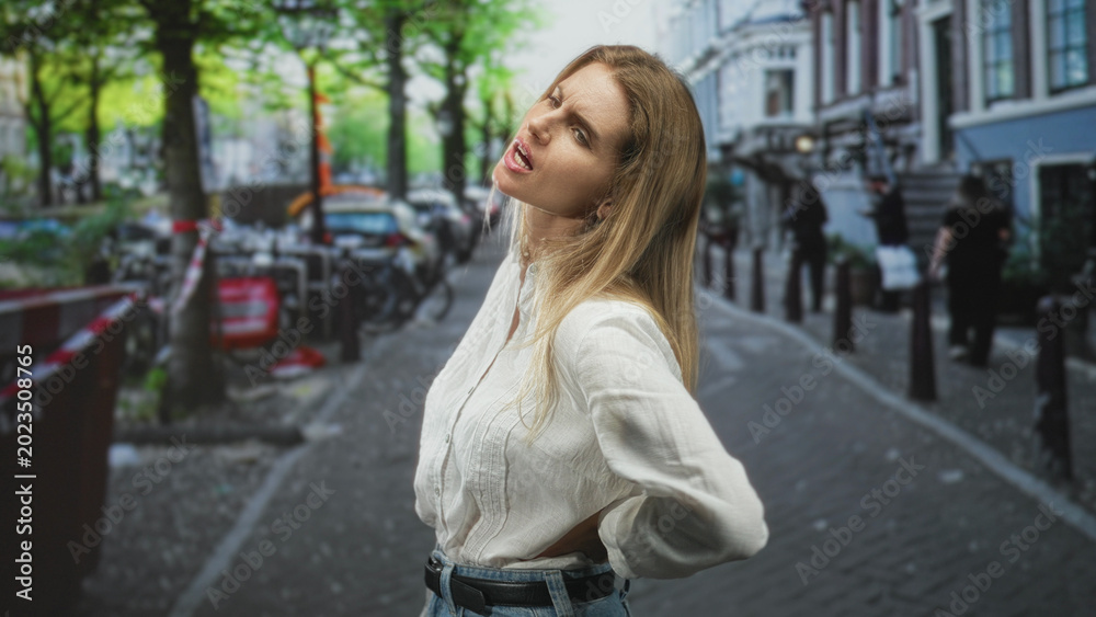 Fototapeta premium Woman in white blouse with hand on lower back for backache on city street amid bicycles and bollards; discomfort everyday strain.
