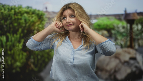 Canvas Print Woman presses fingers into ears in a green park forest, looking upward with annoyed expression; annoyance noise