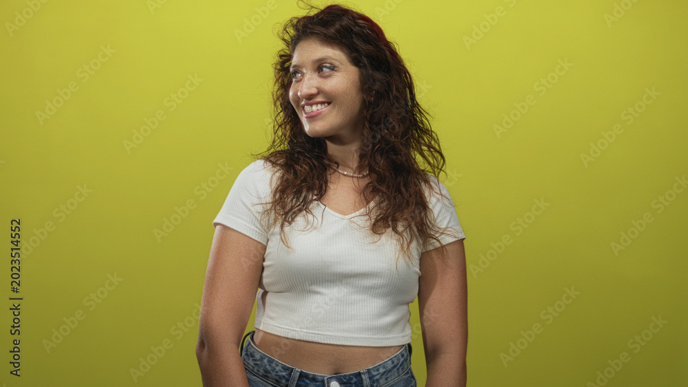 Fototapeta premium Woman smiling with bare midriff wearing white tshirt and jeans three quarter view in lime green studio; confidence casual natural.