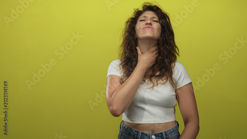 Billede på lærred Young hispanic woman clutching throat with hand, white crop top and jeans showing bare midriff in yellow studio; throat pain