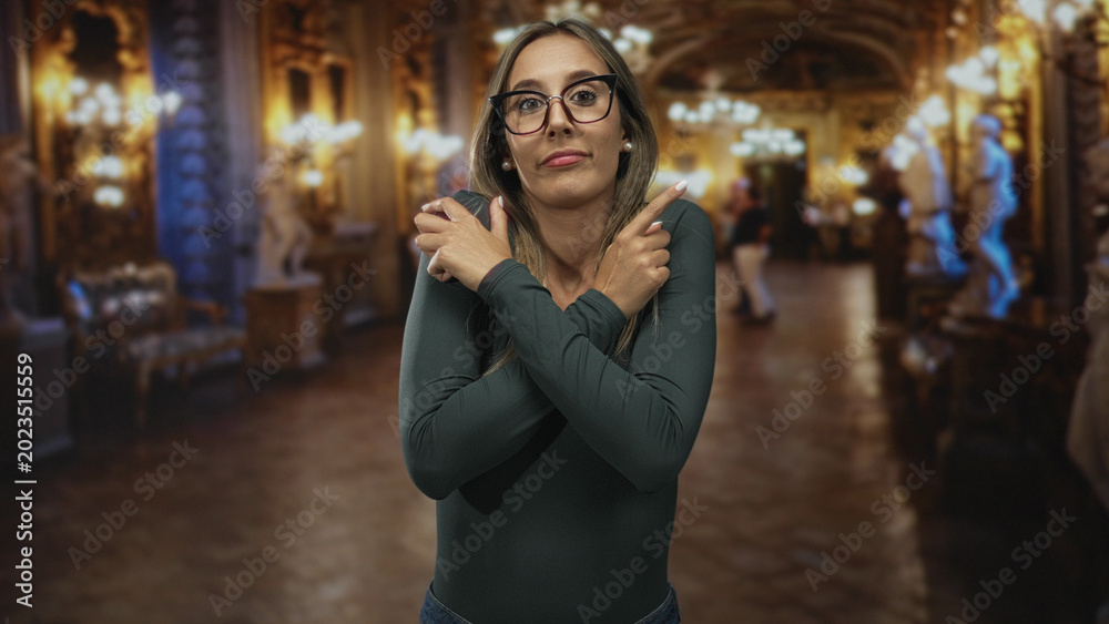 Fototapeta premium Young woman with crossed arms and glasses, wearing pearl earrings and long sleeves, inside ornate museum building; rejection introspection.