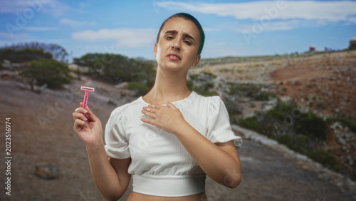 Photos Woman holding pink razor, hand on chest, grimacing on rocky clifftop path with scrubby vegetation and distant sea; disgust hesitation