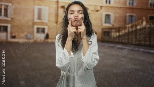 Foto Woman pressing her cheeks with index fingers and puckering lips on a street, wearing a loose white shirt in a sunlit stone plaza; playful expression