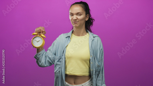 Φωτογραφία Woman holds yellow alarm clock at arm's length with eye roll and annoyed face in studio, yellow crop top and denim overshirt visible; impatience urgency