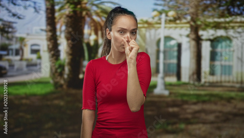Fotografie Woman pinching her nose with fingers in front of a building, wearing a red shirt in an outdoor scene with palm trees and grass; disgust