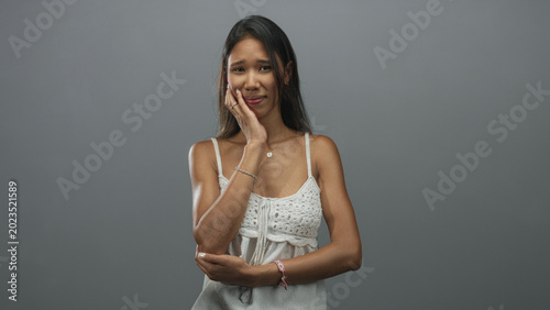 Foto Thai woman pressing cheek with hand and grimacing, wearing white crochet camisole in a studio setting; toothache discomfort