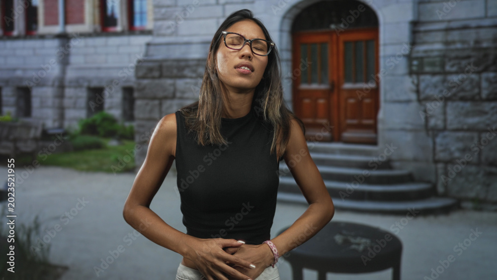 Fototapeta premium Thai woman clutching stomach, grimacing with closed eyes and bent posture showing stomachache in front of stone building steps; discomfort.
