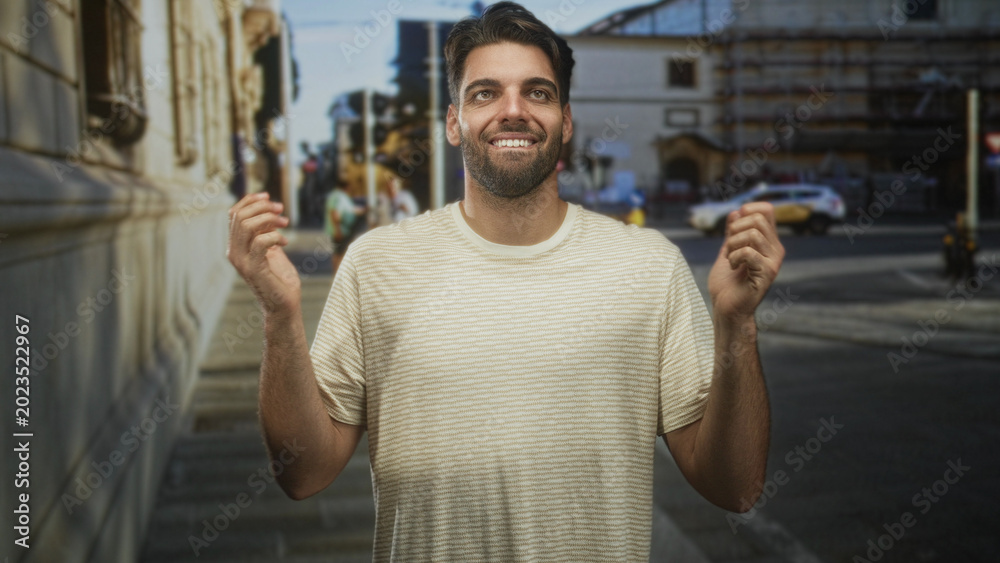 Naklejka premium Hispanic man with beard clapping hands and smiling on a sunlit street beside a building, wearing a striped t shirt while people and a taxi appear behind him; joy gratitude.