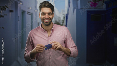 Billede på lærred Man holding a blue creditcard between his hands, smiling and presenting the card on a narrow sunlit street lined with white walls; travel confidence