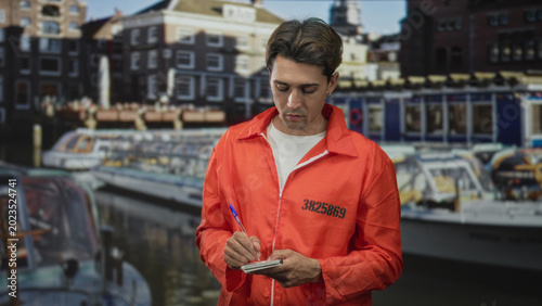 Photos Man in orange inmate jumpsuit writing with a pen on a notepad by canal building and boats; remorse reflection